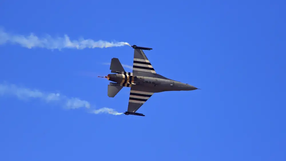 Avion de chasse F-16 en plein vol avec postcombustion allumée et traînées de fumée dans un ciel bleu dégagé.