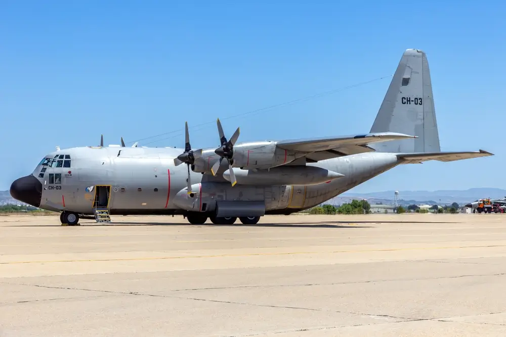 Avion de transport militaire C-130 Hercules stationné sur le tarmac d'une base aérienne, sous un ciel bleu dégagé.