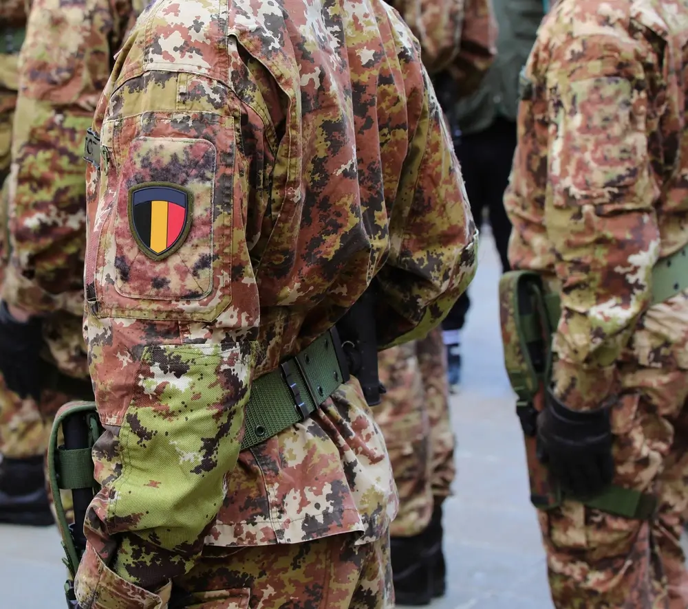Soldat belge en uniforme de camouflage avec écusson national sur le bras et équipement tactique au ceinturon.