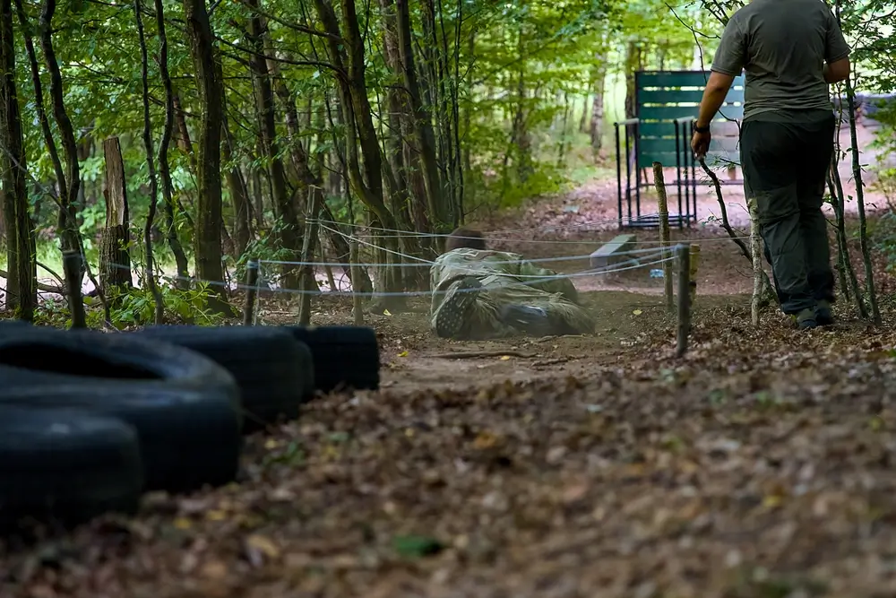 Soldat rampant sous des fils lors d'un test physique militaire sur un parcours d'obstacles en forêt.