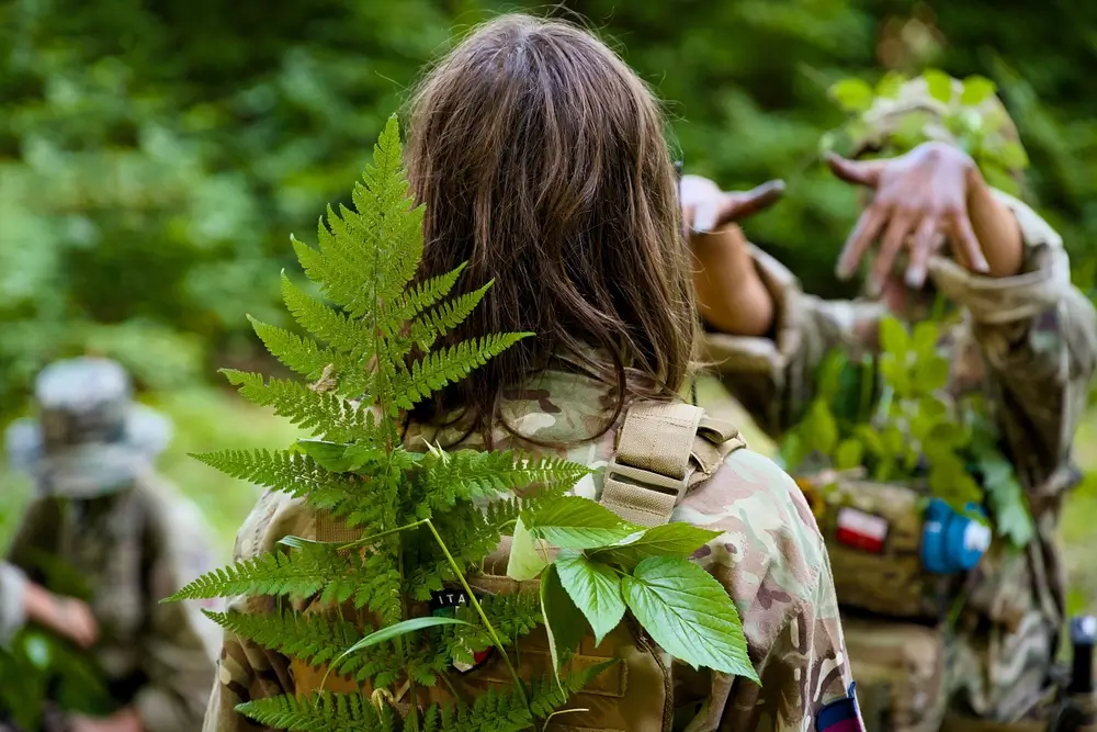 Adolescent en stage militaire utilisant des fougères pour le camouflage lors d'un entraînement en forêt.