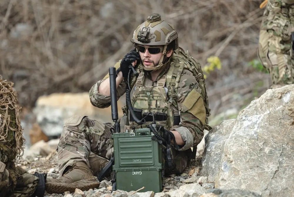 Soldat en tenue de camouflage utilisant un équipement de communication radio tactique sur un terrain rocheux.